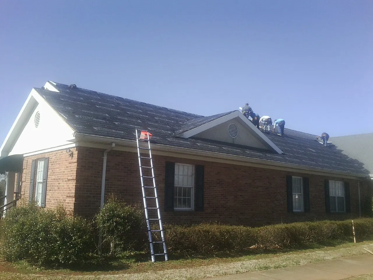 Crew tearing shingles off a brick ranch roof with a ladder set — Metal Roof Repair work by our crew in Ypsilanti